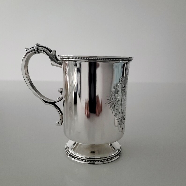 Puede incluir: Una taza de plata con un asa decorativa y un dise&ntilde;o grabado detallado. La taza tiene una base redonda y un acabado liso y pulido.