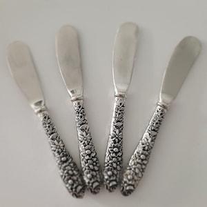 May include: Four silver butter knives with floral patterned handles. The blades are smooth and curved. The handles are intricately detailed with a repeating flower design. The knives are arranged on a white surface.