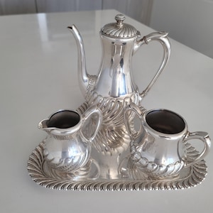 Puede incluir: Un juego de té de plata con tetera, jarra de crema y azucarero. El juego está en una bandeja de plata con un borde decorativo.
