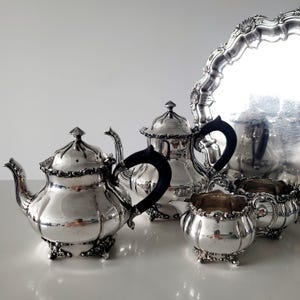 Può includere: Un servizio da tè in argento con vassoio. Il set include una teiera, una lattiera, una zuccheriera e un vassoio. La teiera ha un manico nero e un bordo decorativo. Il set è esposto su una superficie bianca.