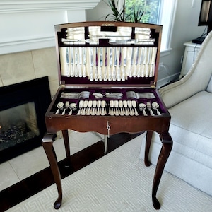 Puede incluir: Un cofre de cubiertos de madera con tapa abatible, abierto para revelar un juego completo de utensilios de plata. El interior está forrado con terciopelo morado. El cofre está sobre cuatro patas curvas y tiene una llave en la cerradura.