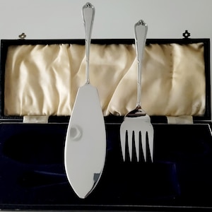 Puede incluir: Un juego de servidor de pastel y tenedor de servir plateados, presentado en una caja de presentación negra con un forro de satén color crema. El servidor tiene una hoja ancha y puntiaguda, y el tenedor tiene cuatro púas. Ambos tienen mangos ornamentados.