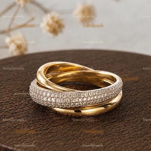 Puede incluir: Un anillo de oro de tres bandas con tres aros entrelazados. Una banda está incrustada con numerosas piedras pequeñas y claras en pavé, mientras que las otras dos bandas son de oro liso y pulido, creando un diseño circular superpuesto y entrelazado.