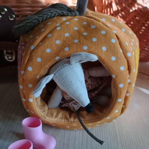May include: A handmade orange fabric pumpkin with white polka dots, housing a gray mouse toy. The pumpkin has a green leaf and gray stem. Two small pink boots are in front of the pumpkin. The scene evokes a cozy, handcrafted aesthetic.