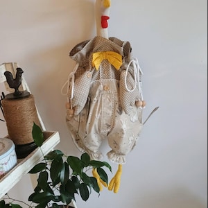 May include: A whimsical, handmade chicken-shaped kitchen accessory. The chicken has a white head, red comb and wattle, and yellow beak and feet. It wears a tan outfit with a yellow bow tie and a polka dot pattern. The overall design is charming and decorative.