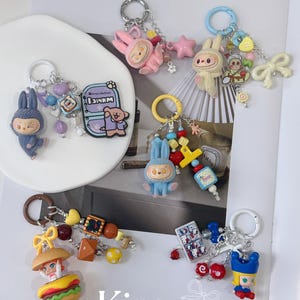 May include: Collection of colorful handmade keychains featuring cartoon characters, food items, and decorative beads. Keychains include a blue bunny, pink bunny, burger, and other charms. The text "Kira.ra Handmade Strap" is visible.