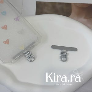 Puede incluir: Una funda transparente para teléfono con diseños de corazones coloridos y herrajes plateados. También hay un accesorio de correa plateado a juego. El texto "Kira.ra Handmade strap" es visible.