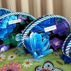 May include: Four half-moon shaped cosmetic bags with a floral pattern in shades of blue, purple, and green. Each bag has a white and blue striped trim and a zipper closure. The bags are labeled "Chubby Cupcake".