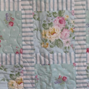 May include: A patchwork quilt featuring floral panels of pink and yellow roses on a pale blue backdrop. The design incorporates white polka dots and sections with blue and white stripes.