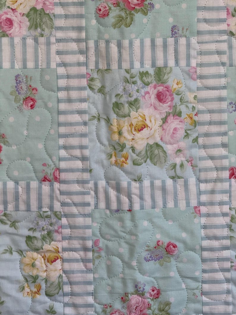 May include: A patchwork quilt featuring floral panels of pink and yellow roses on a pale blue backdrop. The design incorporates white polka dots and sections with blue and white stripes.