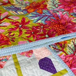 Puede incluir: Un edred&oacute;n colorido con un estampado floral en tonos rosa, naranja y verde. El edred&oacute;n presenta un borde azul claro y un dise&ntilde;o geom&eacute;trico en la esquina con detalles morados y amarillos. El edred&oacute;n est&aacute; hecho de tela.