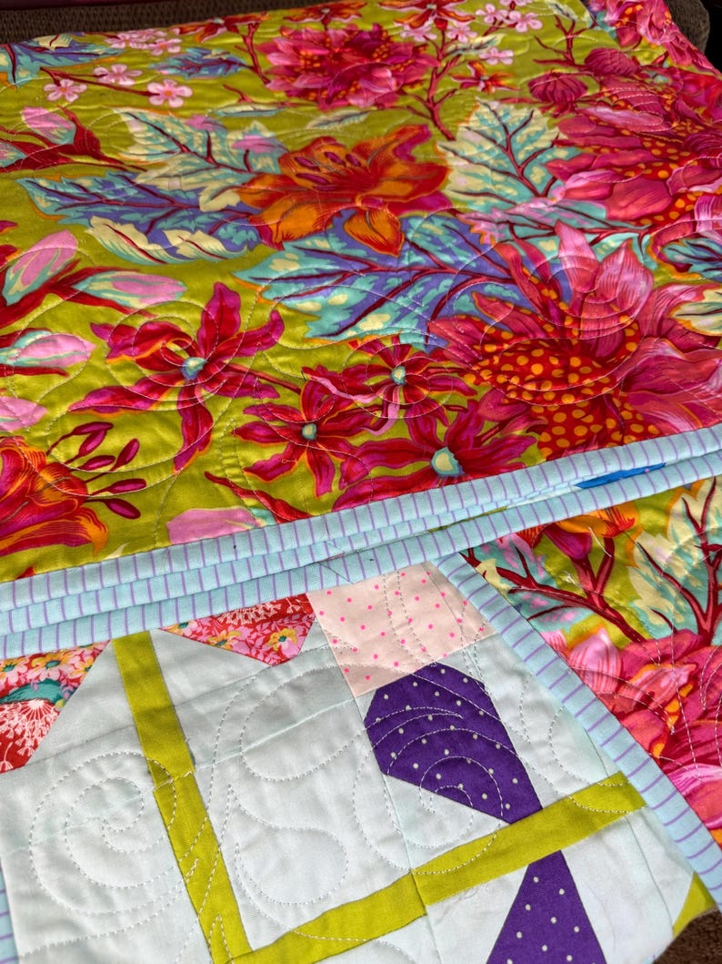 Puede incluir: Un edred&oacute;n colorido con un estampado floral en tonos rosa, naranja y verde. El edred&oacute;n presenta un borde azul claro y un dise&ntilde;o geom&eacute;trico en la esquina con detalles morados y amarillos. El edred&oacute;n est&aacute; hecho de tela.