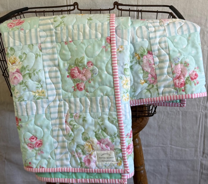 May include: A handmade quilt with a floral and striped pattern in shades of light blue, pink, and white. The quilt has a pink striped border and a label that reads "handmade by Pattie Mamas".