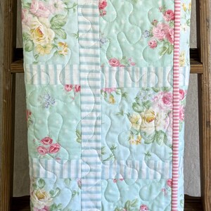 May include: A floral quilt featuring pink and yellow roses on a light blue backdrop. The quilt has a striped border and is displayed on a wooden ladder. The quilt is made of soft fabric and is suitable for a bed or a decorative throw.