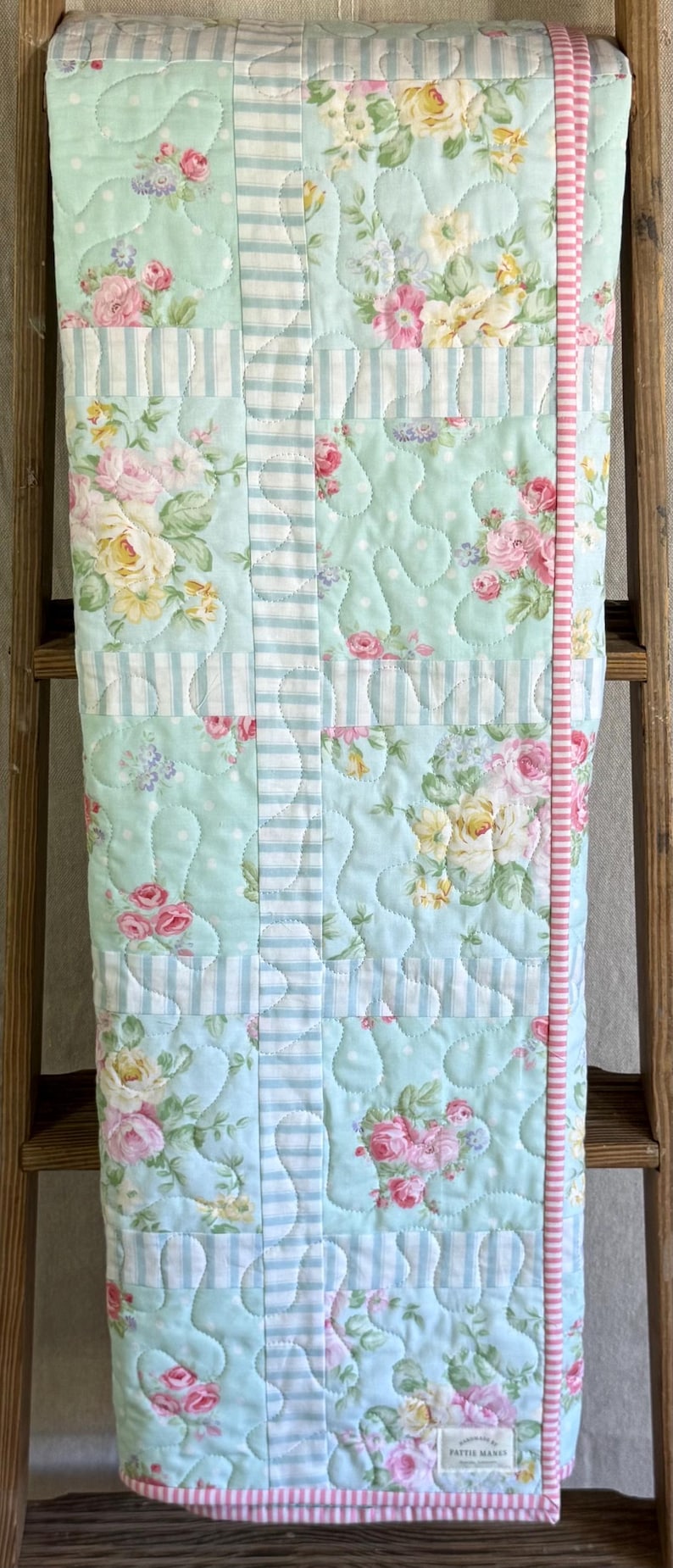 May include: A floral quilt featuring pink and yellow roses on a light blue backdrop. The quilt has a striped border and is displayed on a wooden ladder. The quilt is made of soft fabric and is suitable for a bed or a decorative throw.