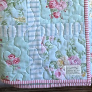 May include: A floral quilted blanket featuring pink roses and green leaves on a pale blue backdrop. It has sections of blue and white stripes and a pink and white striped border. The label reads "handmade by Pattie Manes."