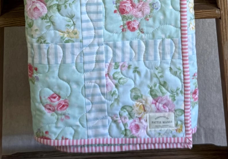 May include: A floral quilted blanket featuring pink roses and green leaves on a pale blue backdrop. It has sections of blue and white stripes and a pink and white striped border. The label reads "handmade by Pattie Manes."