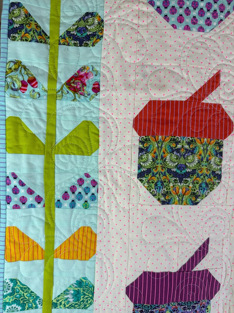 Puede incluir: Primer plano de un edred&oacute;n colorido con un patr&oacute;n floral y geom&eacute;trico. El edred&oacute;n presenta varios patrones de tela, incluyendo florales, a rayas y lunares. La paleta de colores incluye tonos de azul, verde, rojo, amarillo y morado. El edred&oacute;n est&aacute; acolchado a m&aacute;quina con una puntada decorativa.