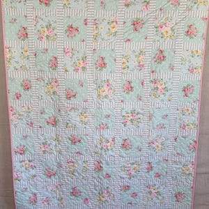 May include: A floral quilt with a pale blue background and a grid pattern. The quilt features squares of pink and yellow roses, with white and pale blue striped borders. The edges are trimmed with a pink border.