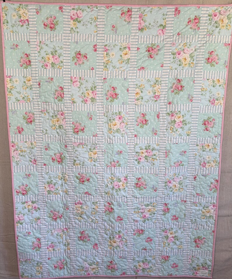 May include: A floral quilt with a pale blue background and a grid pattern. The quilt features squares of pink and yellow roses, with white and pale blue striped borders. The edges are trimmed with a pink border.