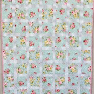 May include: A floral quilt with a pale blue backdrop and a grid design. The quilt showcases squares of pink and yellow roses on a light blue polka dot background, divided by light blue and white striped sections. It has a pink border trim.