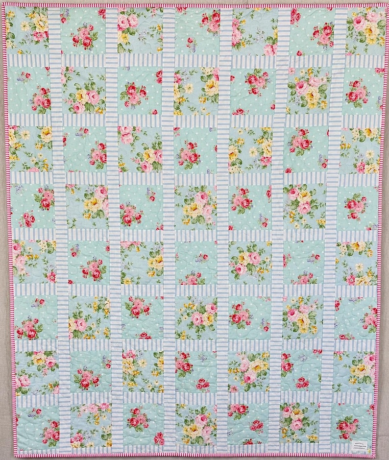 May include: A floral quilt with a pale blue backdrop and a grid design. The quilt showcases squares of pink and yellow roses on a light blue polka dot background, divided by light blue and white striped sections. It has a pink border trim.