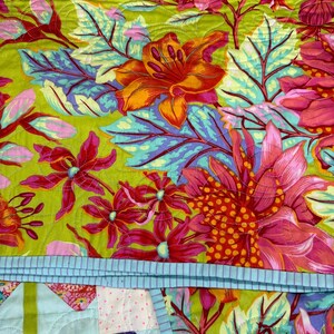 Puede incluir: Primer plano de un edred&oacute;n colorido con un estampado floral. El edred&oacute;n presenta flores grandes en tonos de rosa, naranja y rojo sobre un fondo verde brillante. Las hojas son azules y moradas. Se ve un borde azul claro con rayas.