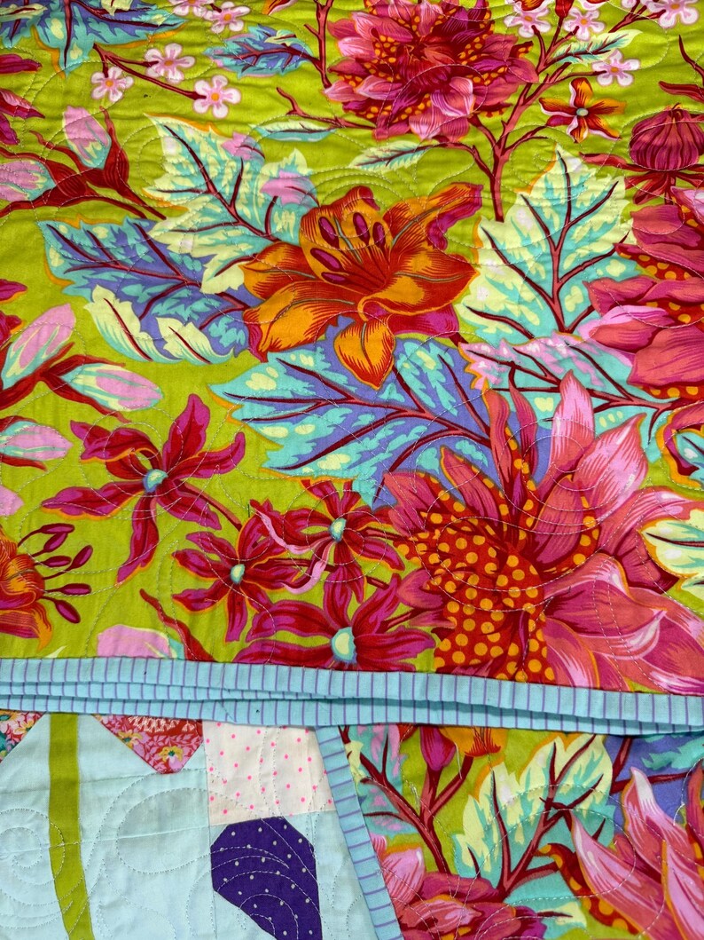 Puede incluir: Primer plano de un edred&oacute;n colorido con un estampado floral. El edred&oacute;n presenta flores grandes en tonos de rosa, naranja y rojo sobre un fondo verde brillante. Las hojas son azules y moradas. Se ve un borde azul claro con rayas.