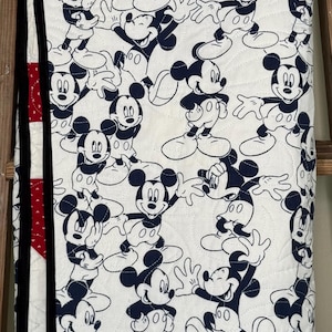 May include: White quilt featuring a repeating pattern of Mickey Mouse in navy blue. The quilt has a red polka dot accent and black trim. The quilt is folded over a wooden ladder.