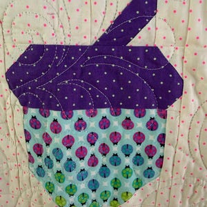 Puede incluir: Un cuadrado de tela acolchada con un dise&ntilde;o de cupcake. La magdalena tiene una parte superior morada con lunares blancos y una base azul claro adornada con patrones de mariquitas. El fondo es blanquecino con lunares rosas. La costura es visible.