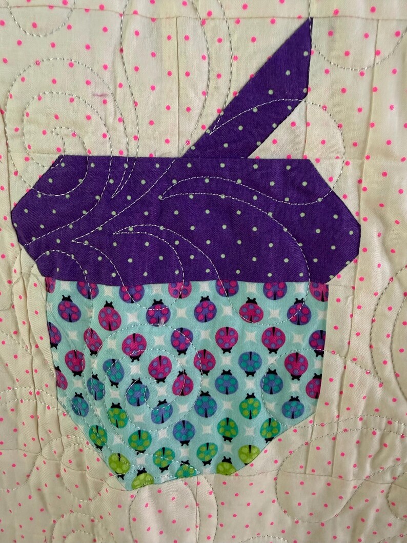 Puede incluir: Un cuadrado de tela acolchada con un dise&ntilde;o de cupcake. La magdalena tiene una parte superior morada con lunares blancos y una base azul claro adornada con patrones de mariquitas. El fondo es blanquecino con lunares rosas. La costura es visible.