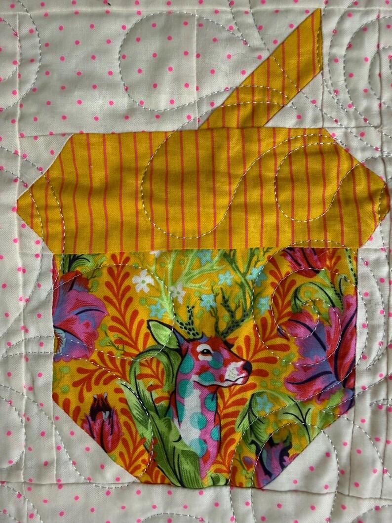 Puede incluir: Un bloque de edred&oacute;n hecho a mano con un ciervo estilizado con manchas y cornamentas coloridas, sobre un vibrante fondo floral. El ciervo est&aacute; encerrado en una forma de taza amarilla con un asa a rayas amarillas. El fondo es blanco con lunares rosas.