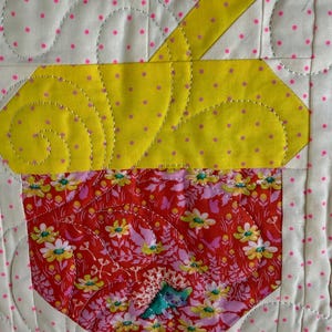 Puede incluir: Un dise&ntilde;o de cupcake acolchado con una parte superior amarilla y una parte inferior floral roja. La magdalena est&aacute; cosida sobre un fondo blanco con lunares rosas. El acolchado presenta l&iacute;neas curvas y una peque&ntilde;a aplicaci&oacute;n azul.