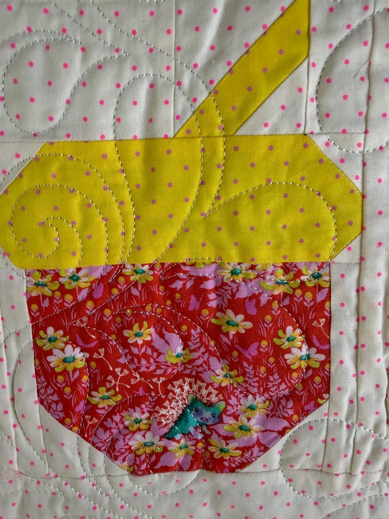 Puede incluir: Un dise&ntilde;o de cupcake acolchado con una parte superior amarilla y una parte inferior floral roja. La magdalena est&aacute; cosida sobre un fondo blanco con lunares rosas. El acolchado presenta l&iacute;neas curvas y una peque&ntilde;a aplicaci&oacute;n azul.