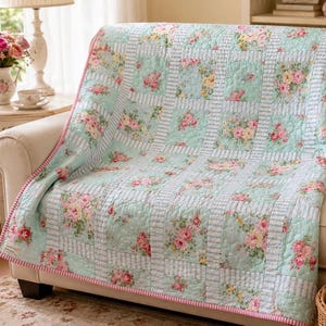 May include: A floral quilt draped over a beige sofa. The quilt features a light blue background with pink and yellow rose bouquets in square patterns. White and blue striped borders and a pink trim complete the design.