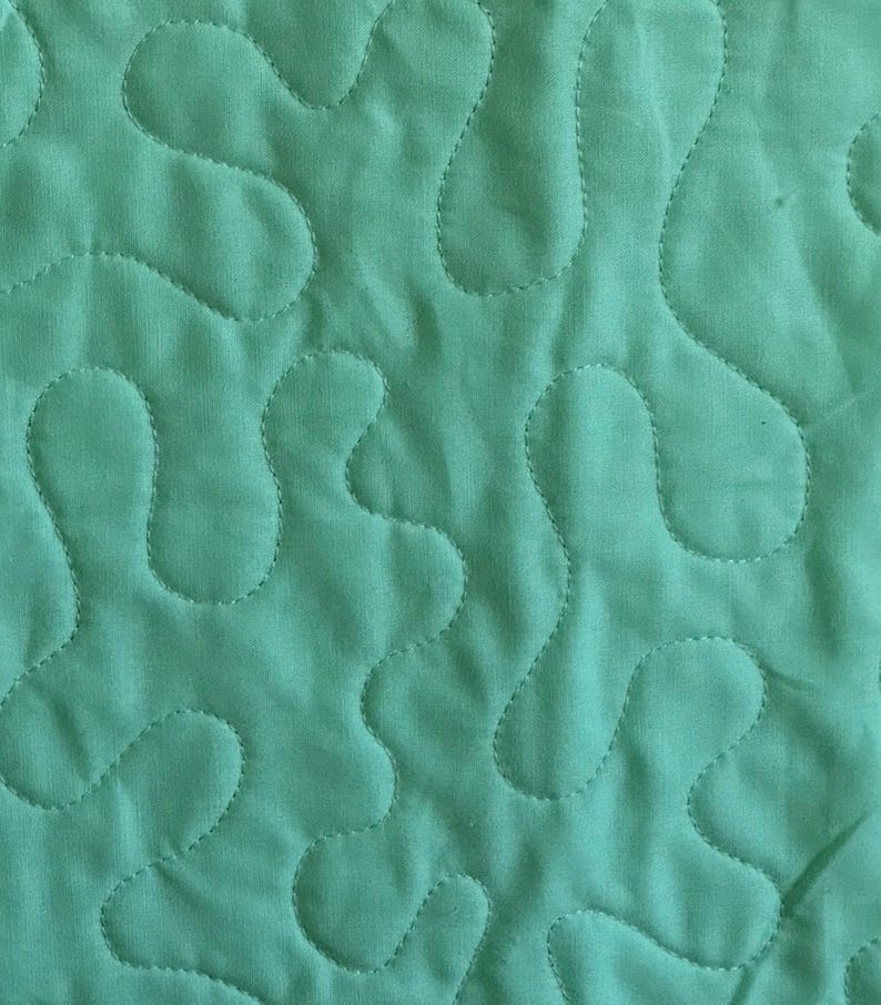May include: Close-up of teal fabric with a quilted design. The quilting pattern features flowing, organic shapes stitched with matching thread. The fabric has a slightly textured appearance, and the stitching creates a subtle, raised effect. The overall aesthetic is soft and inviting.