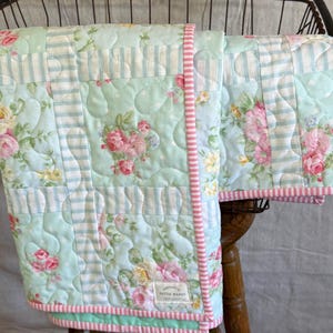 May include: A floral and striped patterned quilt. The quilt has a light blue background with pink and yellow roses, and light blue and white stripes. The edges are trimmed with pink and white striped fabric. The quilt is handmade.