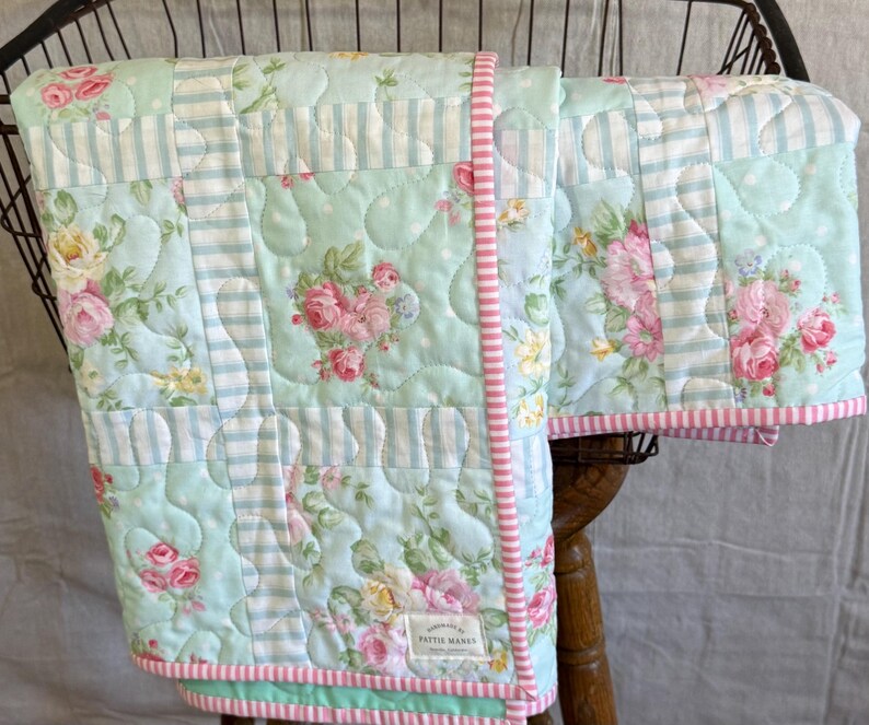 May include: A floral and striped patterned quilt. The quilt has a light blue background with pink and yellow roses, and light blue and white stripes. The edges are trimmed with pink and white striped fabric. The quilt is handmade.