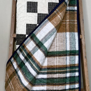 May include: A handmade quilt with a patchwork design. The top features white squares with black and gray plaid accents. The underside has a brown, green, and white plaid pattern. The quilt is trimmed with a navy blue border and is draped over a wooden ladder.