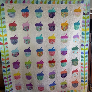 Puede incluir: Un edred&oacute;n hecho a mano con un patr&oacute;n repetido de bellotas coloridas sobre un fondo crema. Las bellotas est&aacute;n hechas de telas estampadas en tonos de azul, verde, morado y rojo. El edred&oacute;n est&aacute; bordeado con dise&ntilde;os florales y geom&eacute;tricos.