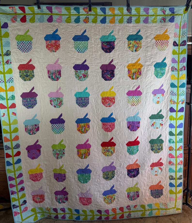 Puede incluir: Un edred&oacute;n hecho a mano con un patr&oacute;n repetido de bellotas coloridas sobre un fondo crema. Las bellotas est&aacute;n hechas de telas estampadas en tonos de azul, verde, morado y rojo. El edred&oacute;n est&aacute; bordeado con dise&ntilde;os florales y geom&eacute;tricos.