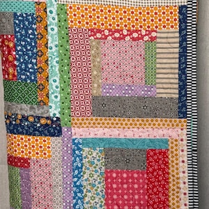 May include: A patchwork quilt featuring a log cabin design with various fabric patterns including floral, geometric, and striped prints in red, blue, green, yellow, and pink. The quilt is finished with a black and white striped border and a small label.