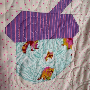 Puede incluir: Un edred&oacute;n hecho a mano con un dise&ntilde;o caprichoso. El edred&oacute;n presenta una forma a rayas moradas y rosas, posiblemente una casa o un &aacute;rbol, sobre un bolsillo con tela de mapache. El fondo es blanco con lunares rosas y costuras decorativas.