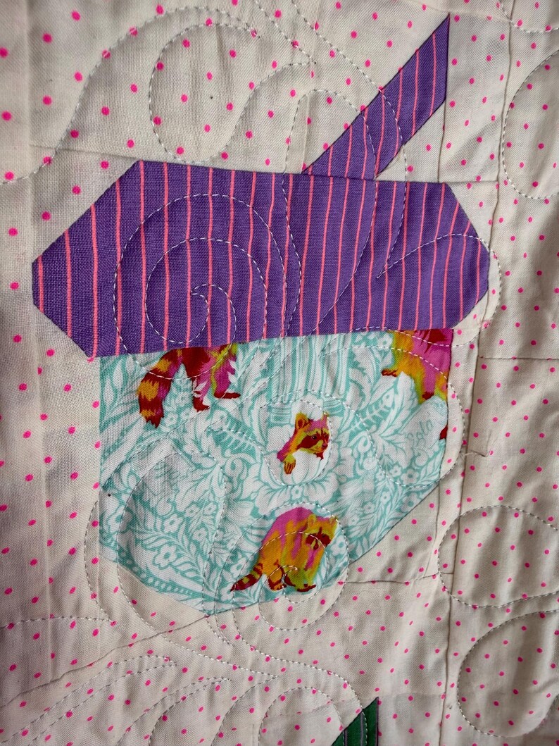 Puede incluir: Un edred&oacute;n hecho a mano con un dise&ntilde;o caprichoso. El edred&oacute;n presenta una forma a rayas moradas y rosas, posiblemente una casa o un &aacute;rbol, sobre un bolsillo con tela de mapache. El fondo es blanco con lunares rosas y costuras decorativas.