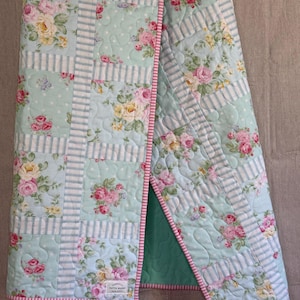 May include: A floral quilt with a light blue and white striped design, featuring pink and yellow roses. The quilt has a pink and white striped border and a green backing. The quilt is folded, revealing the green backing.