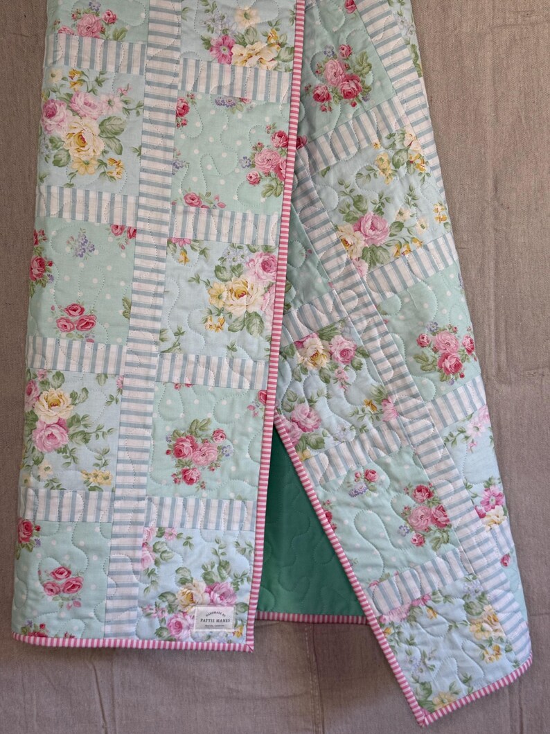 May include: A floral quilt with a light blue and white striped design, featuring pink and yellow roses. The quilt has a pink and white striped border and a green backing. The quilt is folded, revealing the green backing.