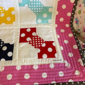 May include: Close-up of a patchwork quilt with a variety of polka dot patterns. The quilt features squares of white, red, yellow, and blue fabric with white polka dots. The border is pink with white polka dots and a gray trim. The text "Chubby Cupcake" is visible.