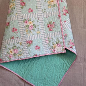 May include: A floral quilt with a light blue backdrop, adorned with pink and yellow roses. It features a striped design and a solid light green reverse with a pink and white striped border. The quilt is folded, displaying the contrasting patterns and colours.