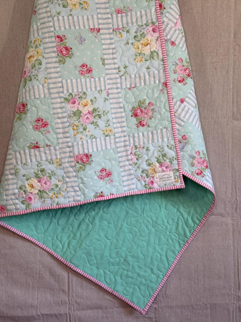 May include: A floral quilt with a light blue backdrop, adorned with pink and yellow roses. It features a striped design and a solid light green reverse with a pink and white striped border. The quilt is folded, displaying the contrasting patterns and colours.