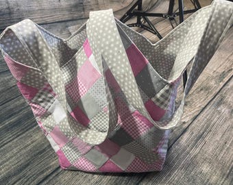 Op de afbeelding: Een grijze en roze patchwork tote bag met twee handvatten. De tas is gemaakt van verschillende stoffen met verschillende patronen, waaronder stippen, ruitjes en ruitjes.